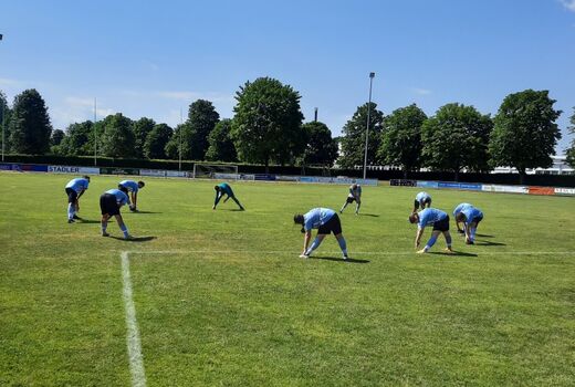 Neun Fußballer in schwarzen Shorts und blauem Trikot stehen im Kreis und machen Dehnungsübungen