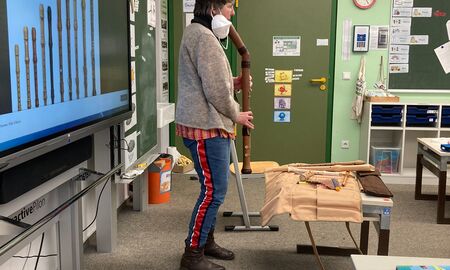 Die Musiklehrerin steht vor der Tafel mit Bildern von verschiedenen Holzflöten und bläst in ein großes Holzblasinstrument.