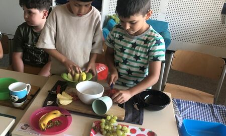 Ein Erstklässler sitzt an einem Tisch. Zwei weitere Erstklässler stehen an einem Tisch und schneiden Obst.