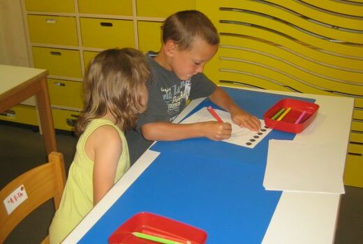 Ein Junge schreibt auf einem Arbeitsblatt den Buchstaben O nach, ein Mädchen schaut zu.