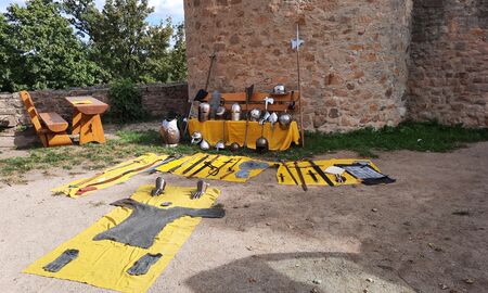 Ritterverkleidung und Requisiten wie Kettenhemd, Helme, Schwerter und weitere Waffen liegen vor einer Burgmauer auf gelben Tüchern ausgebreitet.