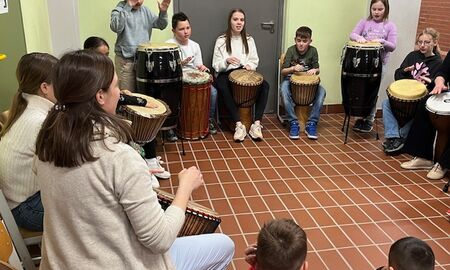 In einem großen Kreis stehen und sitzen viele Kinder an unterschiedlichen Trommeln. Eine Lehrerin macht den Rhythmus an einer Trommel vor. Die Kindern trommeln mit oder nach.