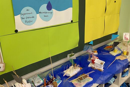 Unter einer Tafel mit dem bisherigen Stundenverlauf stehen auf einem blauen Tuch die fertigen Wasserfahrzeuge.