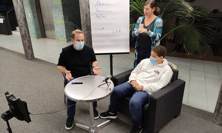 Herr Ulrich stellt dem Schüler Fragen. Beide sitzten nebeneinander und haben eine Maske auf. Im Hintergrund steht Frau Edelmann und dolmetscht.