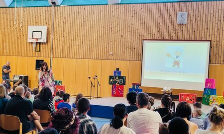 Die Schulleiterin steht vorne neben der Bühne und hält eine Rede. Auf der Leinwand sieht man eine Clipart von einem Bäcker mit der Unterschrift "der Bäcker / die Bäckerin". Im Publikum sitzen die neuen Erstklässler und Erstklässlerinnen mit ihren Eltern.