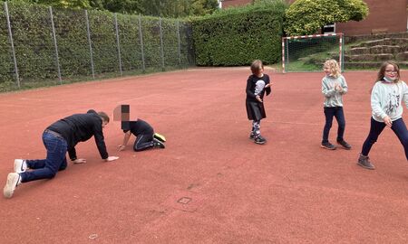 Drei Zweitklässlerinnen laufen in einer Reihe hintereinander rechts aus dem Bild. Links im Bild krabbeln der Musiklehrer und ein Schüler auf allen Vieren aufeinander zu.