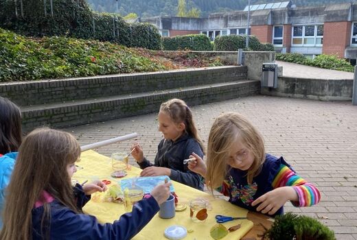 Wir basteln mit unseren Fundstücken aus dem Wald herbstliche Teelichtgläser. Vier Kinder sitzen an einer Bank im Freien und basteln mit Moos und Blättern.