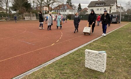 Ein Betonseitenteil wird von einem Jugendlichen auf einer Sackkarre transportiert. Viele Menschen sind auf dem Schulhof