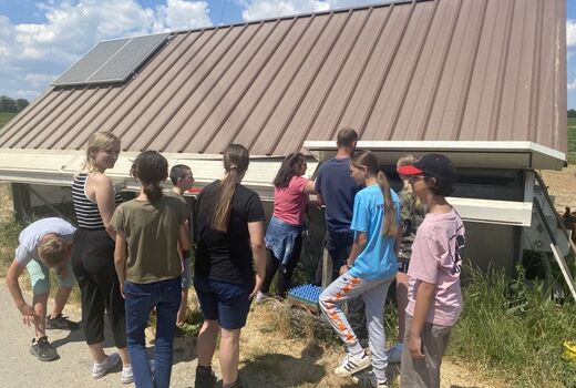 Acht SchülerInnen stehen vor dem Hühnerstall. Im Hintergrund holen zwei Mädchen und ein Mann Eier aus dem Legenest.