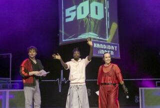 Drei ModeratorInnen stehen auf einer lilafarben beleuchteten Bühne. Zentral hinter ihnen ist ein großes Schild mit der Aufschrift Kandidat:innen und 500.