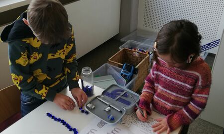 Ein Kind legt mit blauen Muggelsteinen den Buchstaben M. Daneben arbeitet ein Kind an einem Arbeitsblatt.
