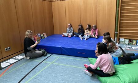 Eine Lehrerin stellt einer Gruppe Kinder, die auf einer Matte in einer Turnhalle sitzen, Bücher vor