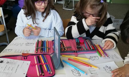 Mehrere Kinder sitzen gemeinsam an einem Tisch. Sie schreiben mit Buntstiften den Groß- und Kleinbuchstaben M bunt. Im Hintergrund sitzen weitere Kinder an ihren Tischen.