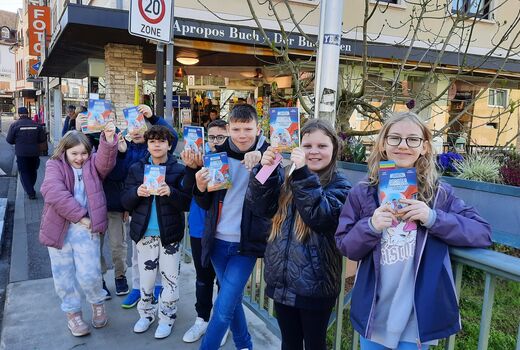 Die Schülerinnen und Schüler stehen vor der Buchhandlung auf einer Brücke und halten ihr neues Buch in den Händen.