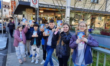 Die Schülerinnen und Schüler stehen vor der Buchhandlung auf einer Brücke und halten ihr neues Buch in den Händen.