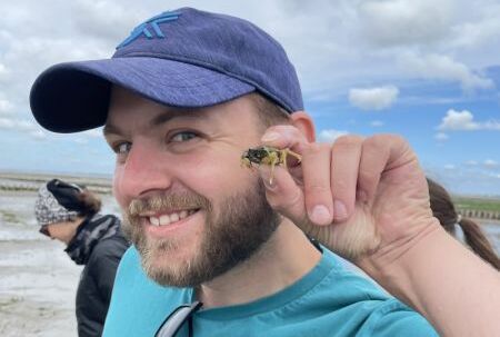 Ein Mann mit Schirmmütze lacht in die Kamera. Er hält eine kleine Krabbe in die Höhe.