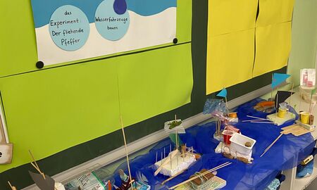 Unter einer Tafel mit dem bisherigen Stundenverlauf stehen auf einem blauen Tuch die fertigen Wasserfahrzeuge.