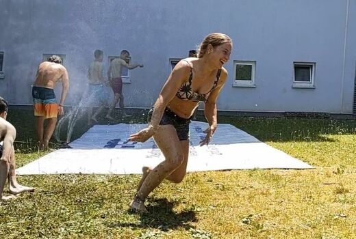 Eine LKW-Plane wird mit Wasser besprenkelt. Mehrere Personen befinden sich um die Plane herum. Eine Frau läuft lachend nach vorne.