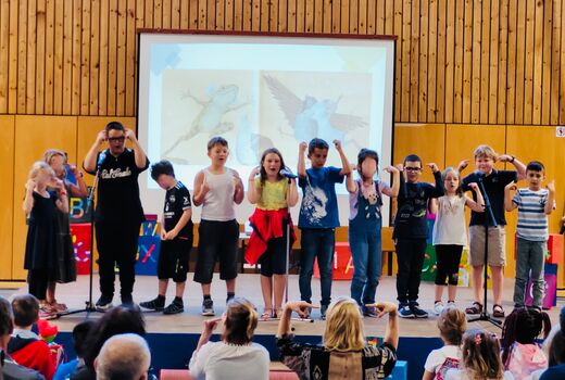 Zwölf Schüler und Schülerinnen stehen in einer Reihe auf der Bühne und führen etwas mit Bewegungen vor.