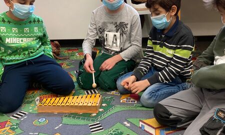 Vier Zweitklässler sitzen auf einem Spielzeugteppich und ein Junge spielt mit einem Holzschlägel auf einem Glockenspiel. Die anderen Kinder schauen ihm dabei zu.