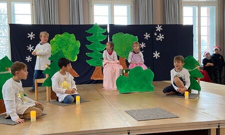 Zwei Mädchen in schönen Kleidern sitzen auf Stühlen. Zwei Jungen mit Sternen auf der Brust tragen Lichter