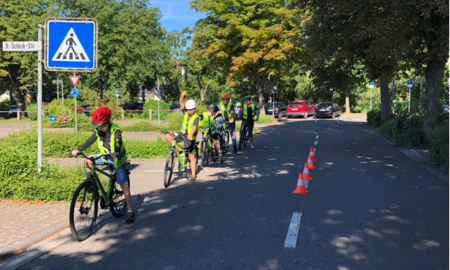 Sechs Kinder in Warnwesten und Helmen warten auf Fahrrädern auf einer Übungsstraße in einer Reihe hintereinander an einem Zebrastreifen.