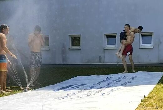 Auf dem Rasen liegt eine LKW-Plane. Seitlich läuft ein Wassersprinkler. Vier Personen stehen um die Plane.