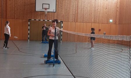 Zu sehen ist eine Erzieherin die an der Seite des Netzes steht. Im Hintergrund sind zwei Spielerinnen zu sehen.