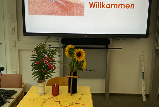 Altar mit Kerze, Blumen und rotem Faden. Im Hintergrund am Smartboard ein Begrüßungsbild mit den Worten "Herzlich Willkommen"