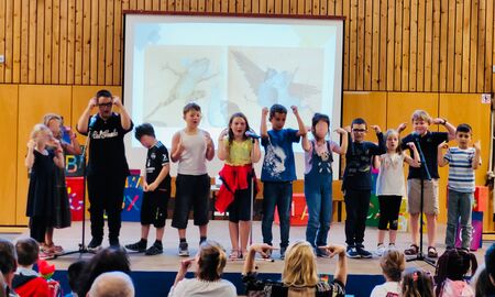 Zwölf Schüler und Schülerinnen stehen in einer Reihe auf der Bühne und führen etwas mit Bewegungen vor.