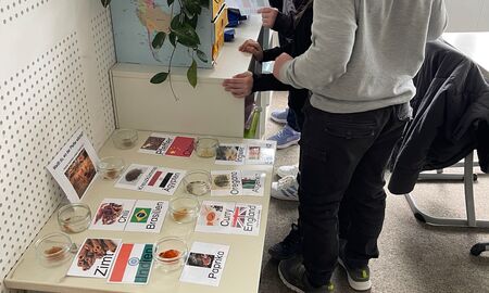 Vorne steht ein Tisch mit verschiedenen Gewürzen aus unterschiedlichen Ländern. Dort stehen Schüsseln mit Gewürzen, Schilder mit der Bezeichnung, Ländernamen und Flaggen. Drei Kinder stehen an dem Regal dahinter und haben ein Schild einer Flagge.