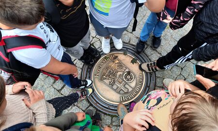 Kinder stehen im Kreis um einen verzierten Gullideckel mit dem Schriftzug Kreisstadt Heppenheim und einem Wappen herum. Das Foto zeigt von oben die Füße der Kinder um den Deckel herum.