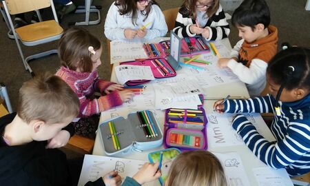 Mehrere Kinder sitzen gemeinsam an Tischen. Sie schreiben auf einem Arbeitsblatt mit Buntstiften den Groß- und Kleinbuchstaben M bunt. Sie malen die Maus auf dem Arbeitsblatt an.