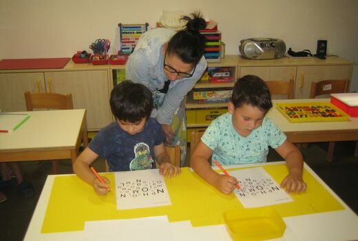 Zwei Jungen suchen und umkreisen auf einem Arbeitsblatt den Buchstaben O. Eine Lehrerin steht dahinter und beobachtet sie.