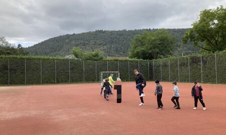 Die Musiklehrerin läuft ganz vorne, ihr folgen acht Kinder und der Musiklehrer. Alle laufen mit den Füßen rhythmisch auftretend in einer Reihe hintereinander über den Sportplatz.