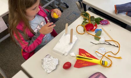 Eine Erstklässlerin sitzt an ihrem Tisch, auf dem ihr Mäppchen und viele Utensilien verstreut liegen, und baut ihr Wasserfahrzeug.