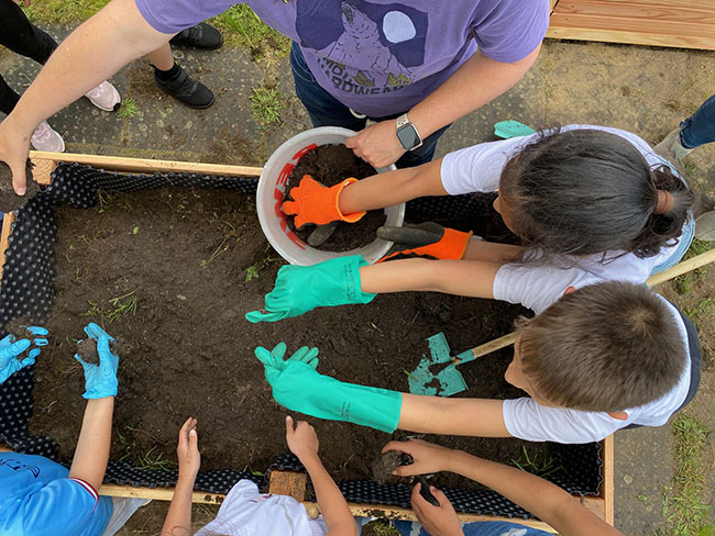 Die Grundschülerinnen und Grundschüler füllen das Hochbeet mit Erde.